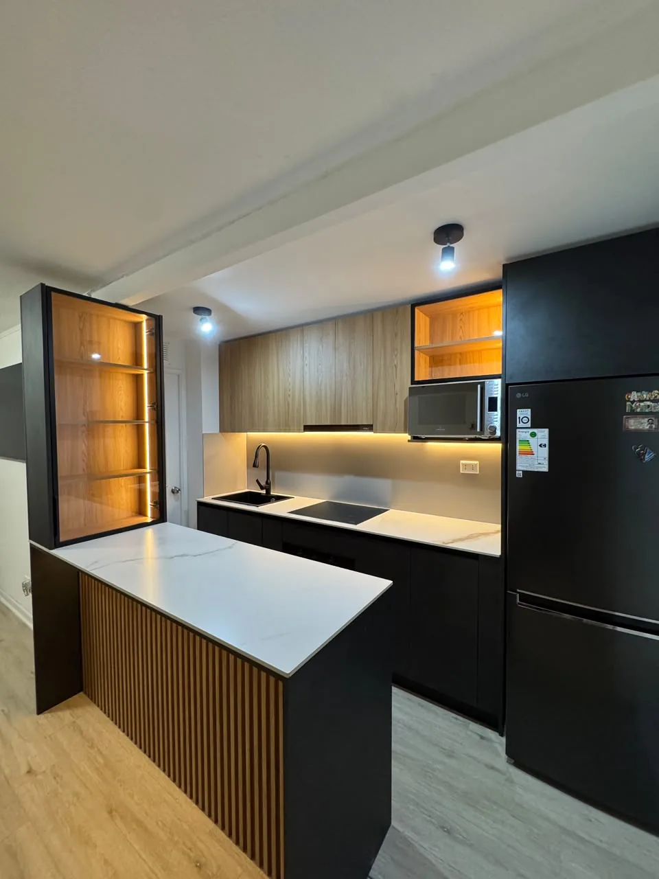 Cocina a medida con muebles negros y detalles en madera, isla con cubierta de porcelánico blanco y vitrina con iluminación LED, en Rancagua